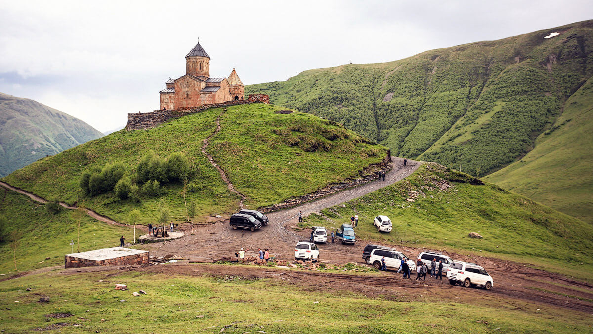У подножья горы стоят таксисты и автомобили жителей Казбеги